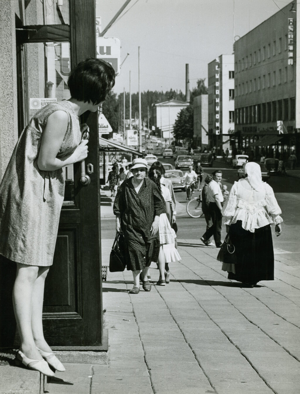 Vanha mustavalkoinen valokuva Riihimäen Hämeenkadulta, jossa nainen katsoo romani-naisen perään kaupan ovelta.